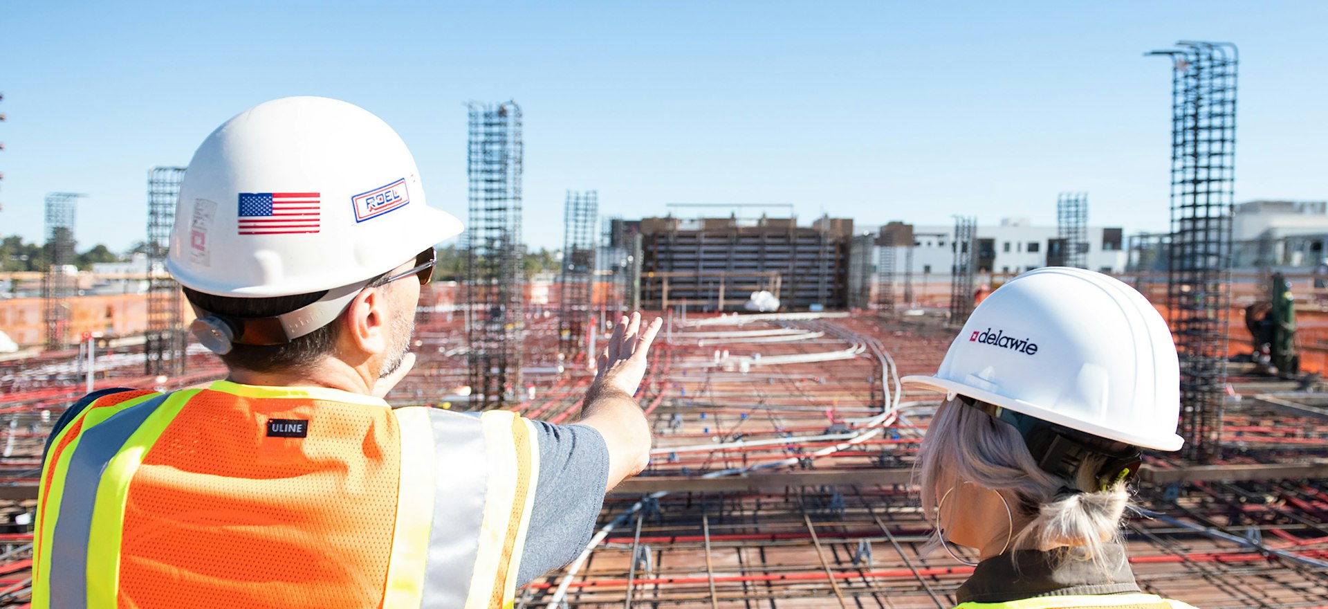 Two construction professionals in branded hard hats and hi-vis vests on the rebar deck of a commercial build, one pointing out the work ahead, the kind of trade business the free AI visibility check is run for