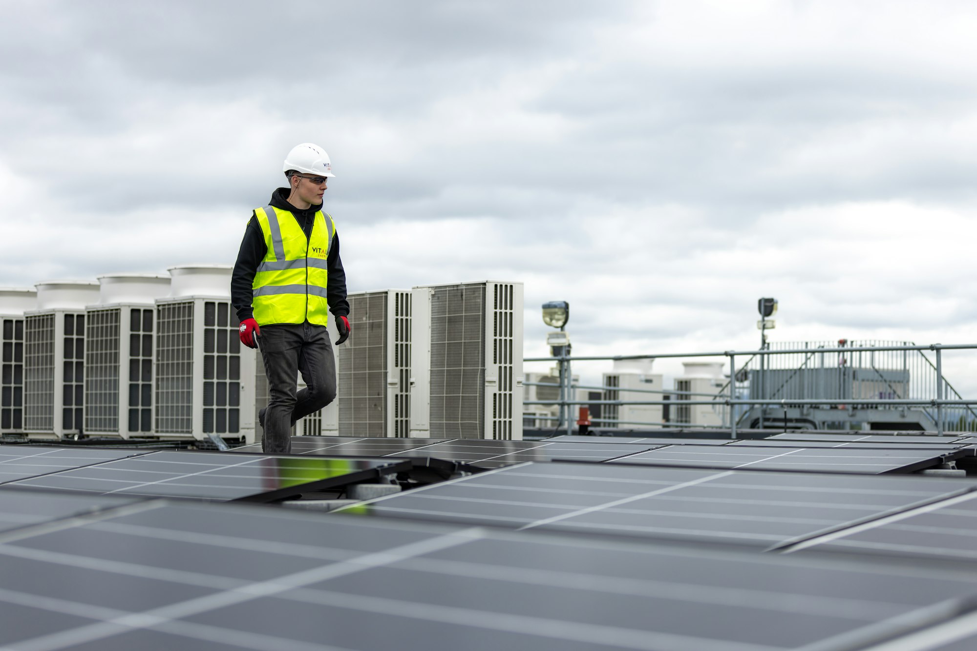 A worker in hi-vis walking across a large flat commercial roof past rooftop plant and solar panels