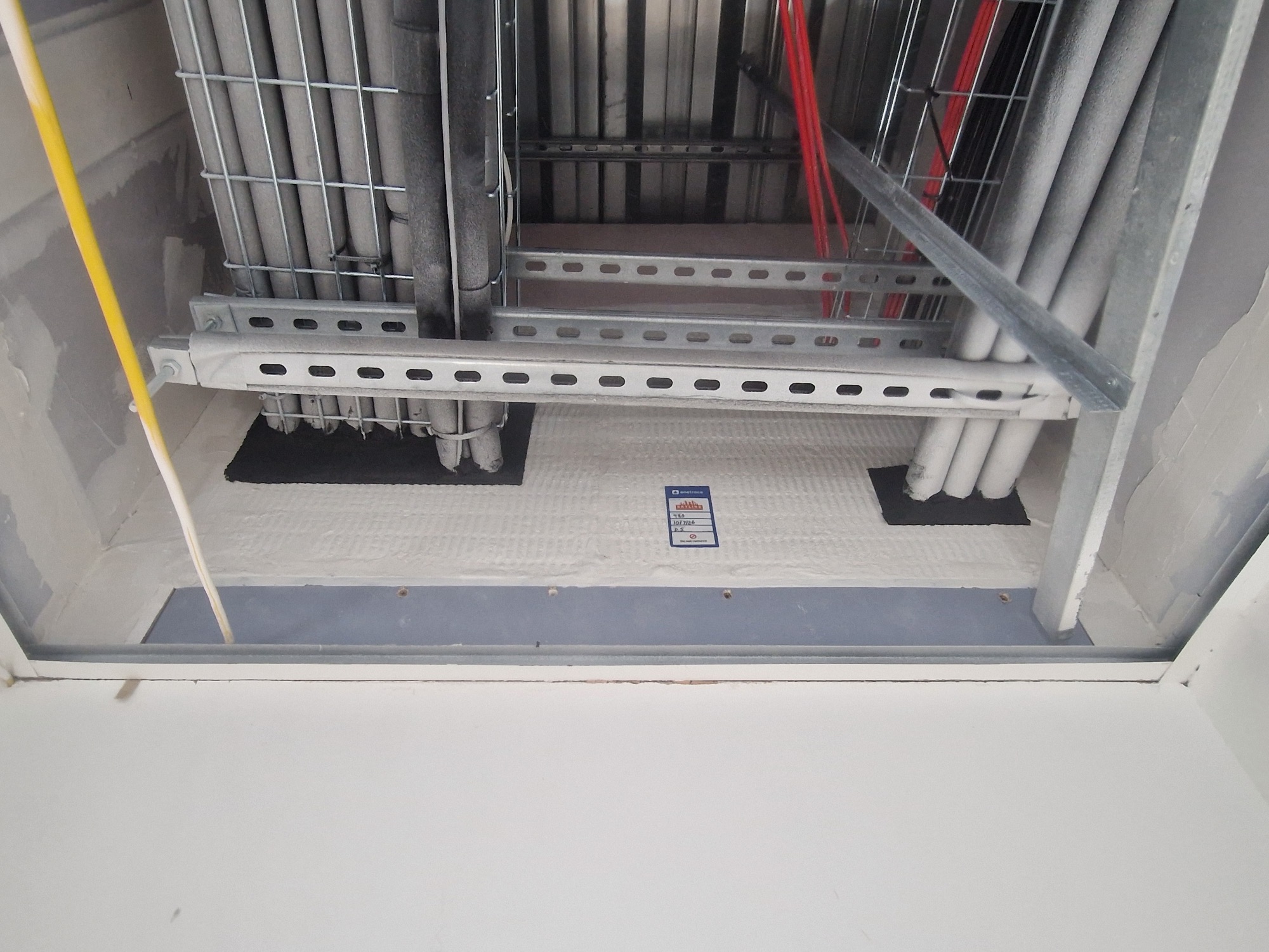 Fire compartmentation held at a compartment floor: pipes and cables cross a riser shaft through a tested firestopping seal with coated batts, each seal carrying its own blue installation tag