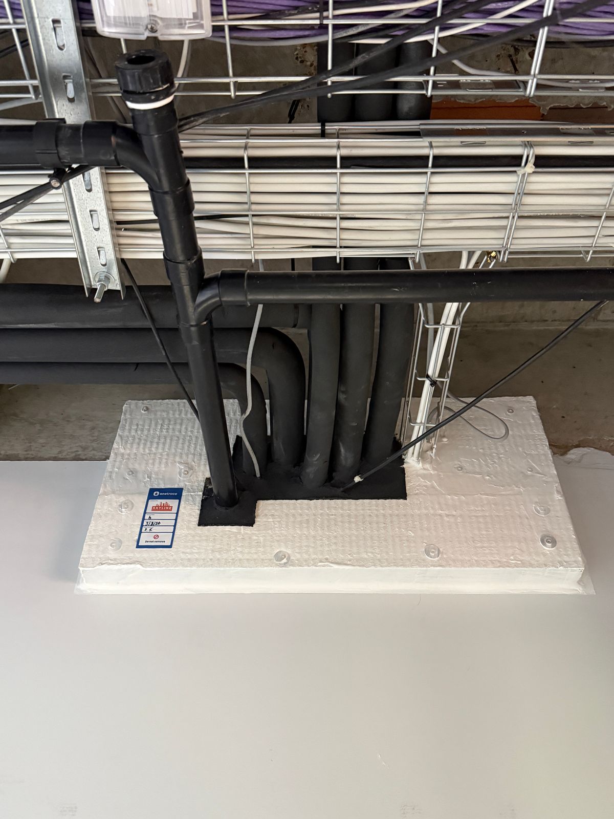A firestopping seal around black pipes and cables, carrying a blue onetrace inspection tag